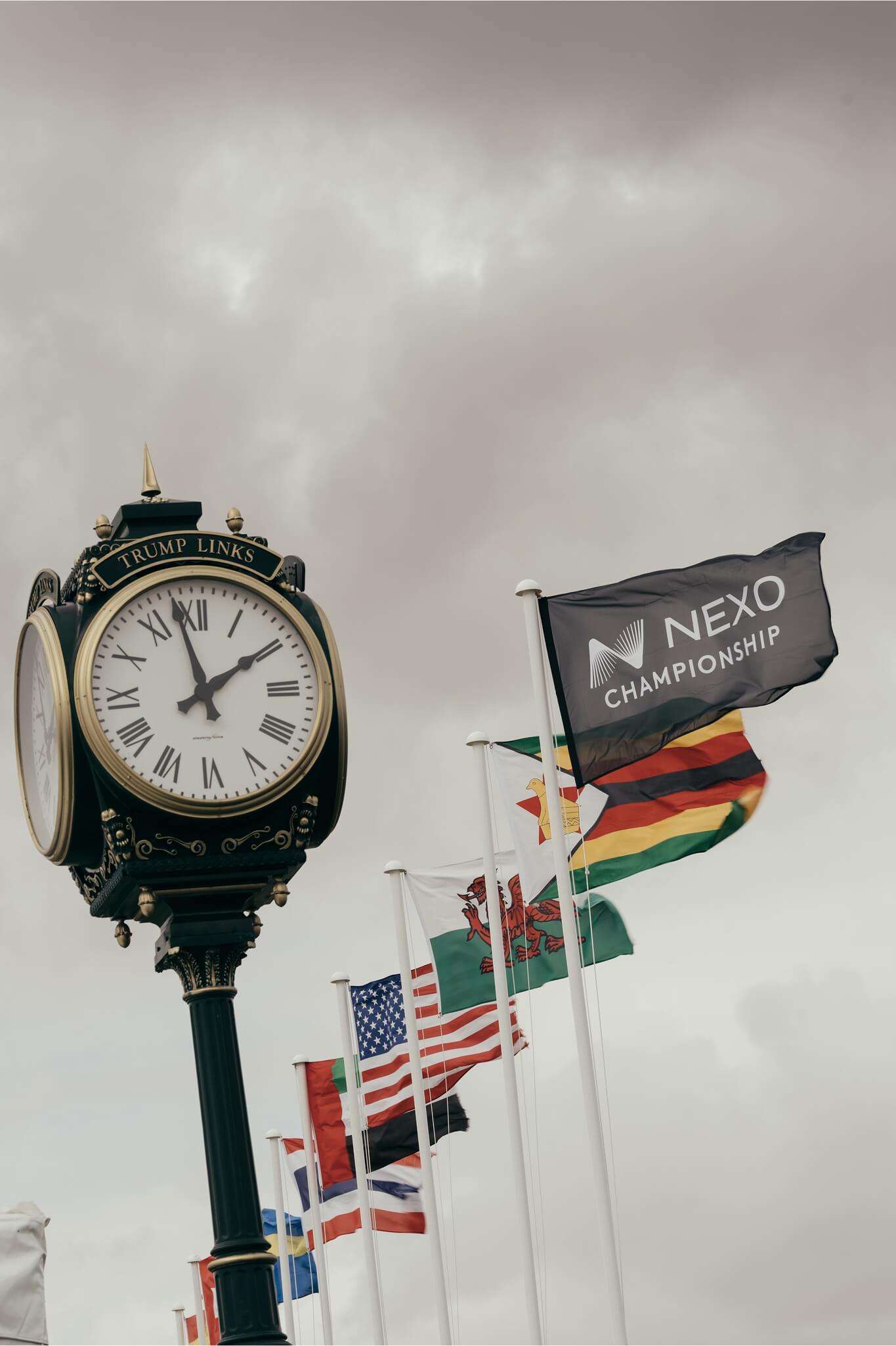 A decorative clock labeled “Trump Links” stands beside several national flags and a Nexo Championship flag waving in the wind.