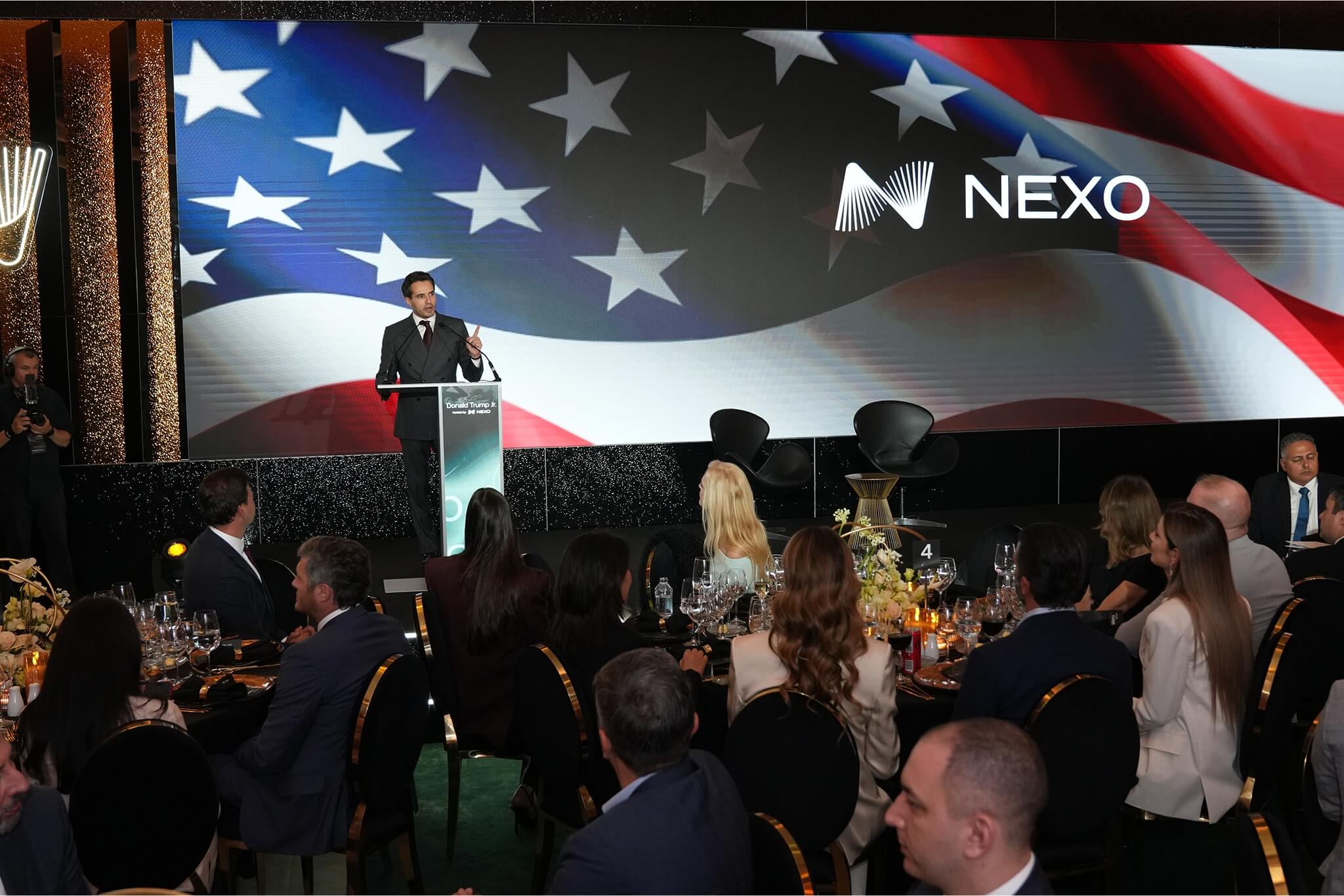 Antoni Trenchev at a Nexo-branded gala event stands at a podium with the American flag projected behind him, addressing a seated audience at elegantly set dinner tables.